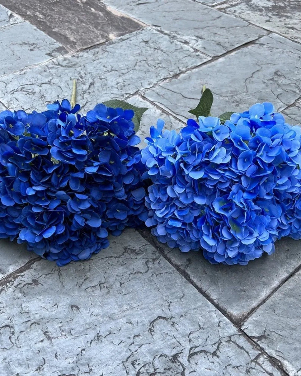 Two blue artificial hydrangea bunches side by side on paving; each order is one bunch