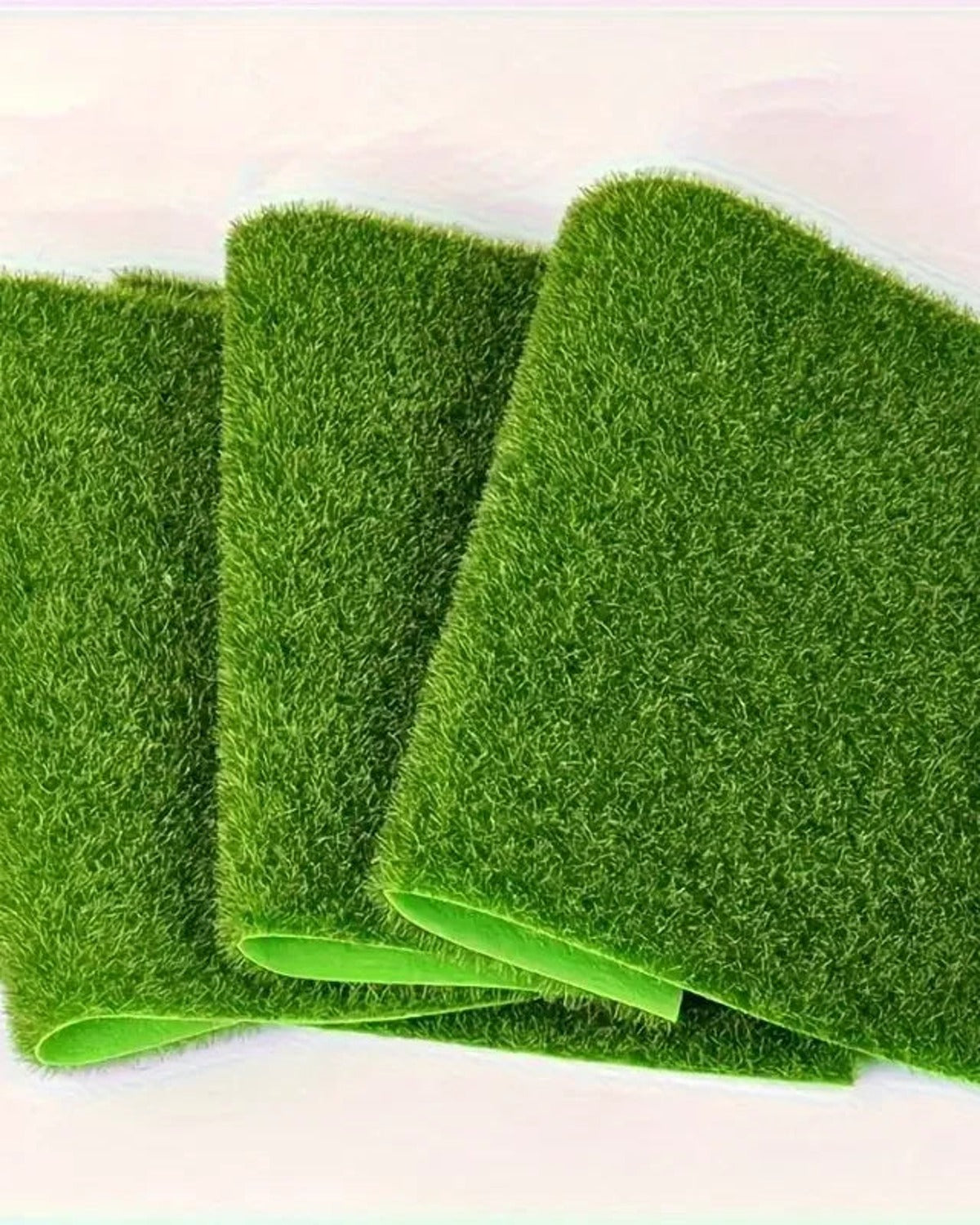 Three artificial grass mats standing on edge, showing the flexible fabric backing; each order is one mat