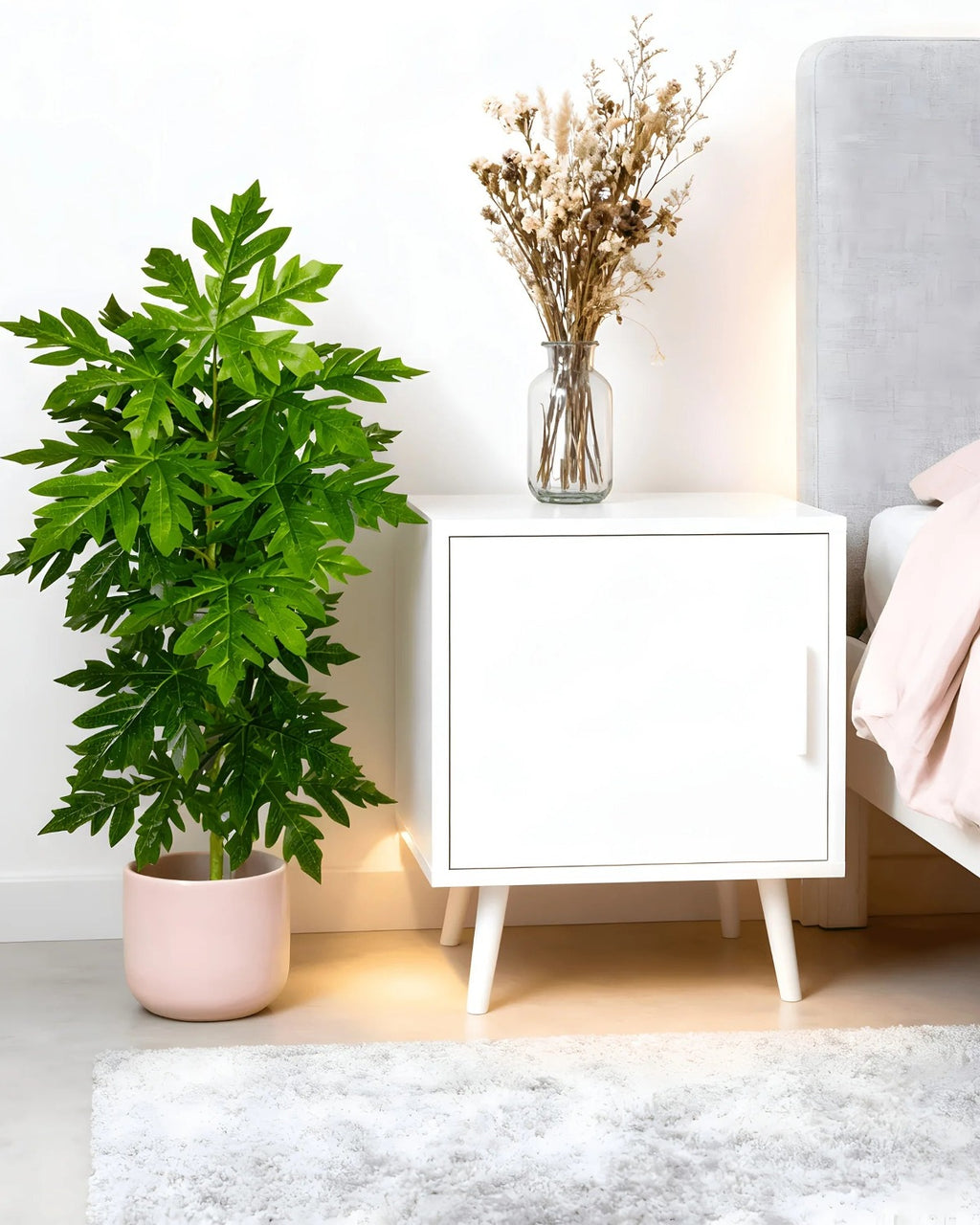 An artificial papaya leaf plant beside a bedside cabinet in a bedroom; pot not included