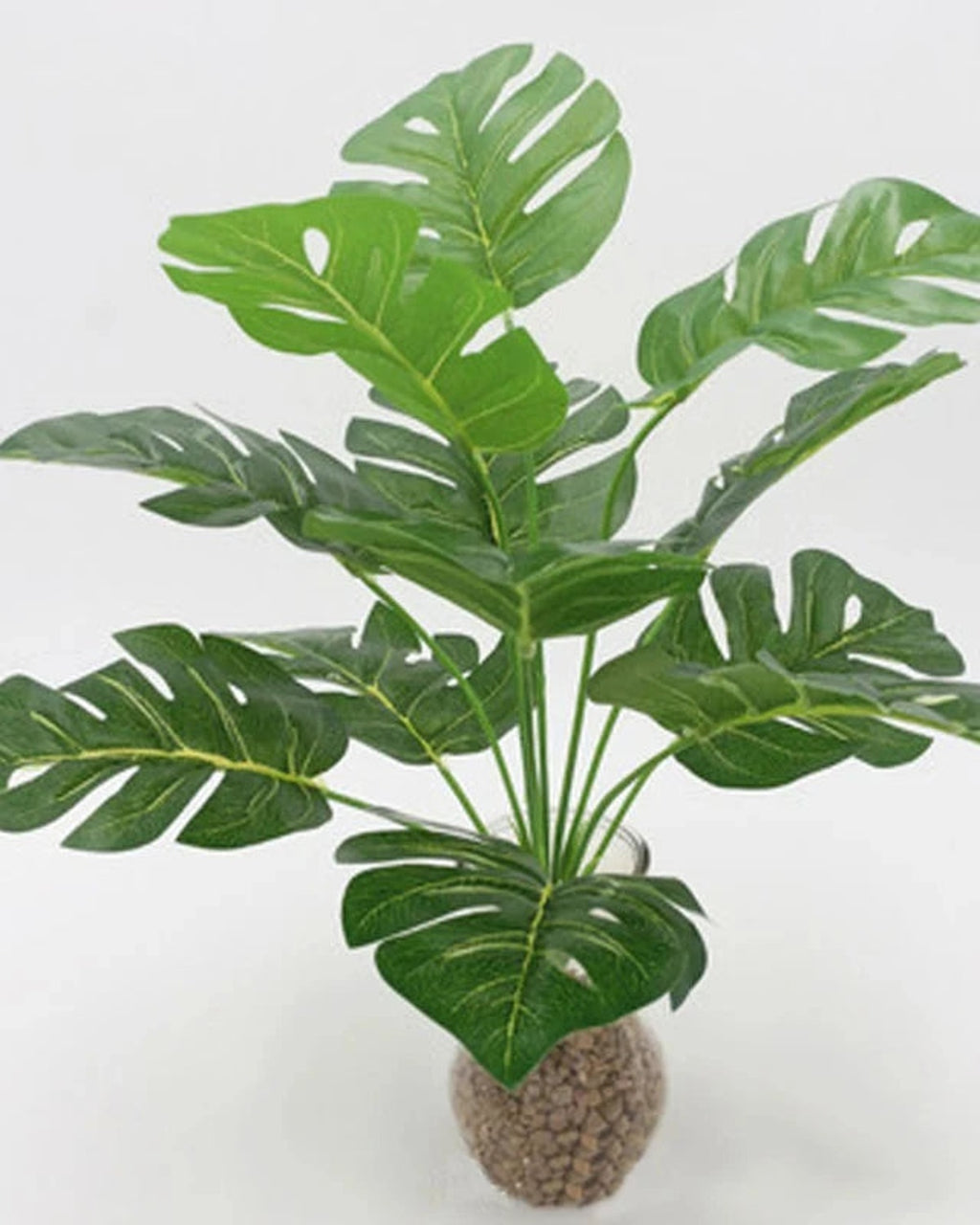 An artificial monstera plant with its stems gathered above a bed of gravel; pot not included