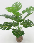 An artificial monstera plant with its stems gathered above a bed of gravel; pot not included