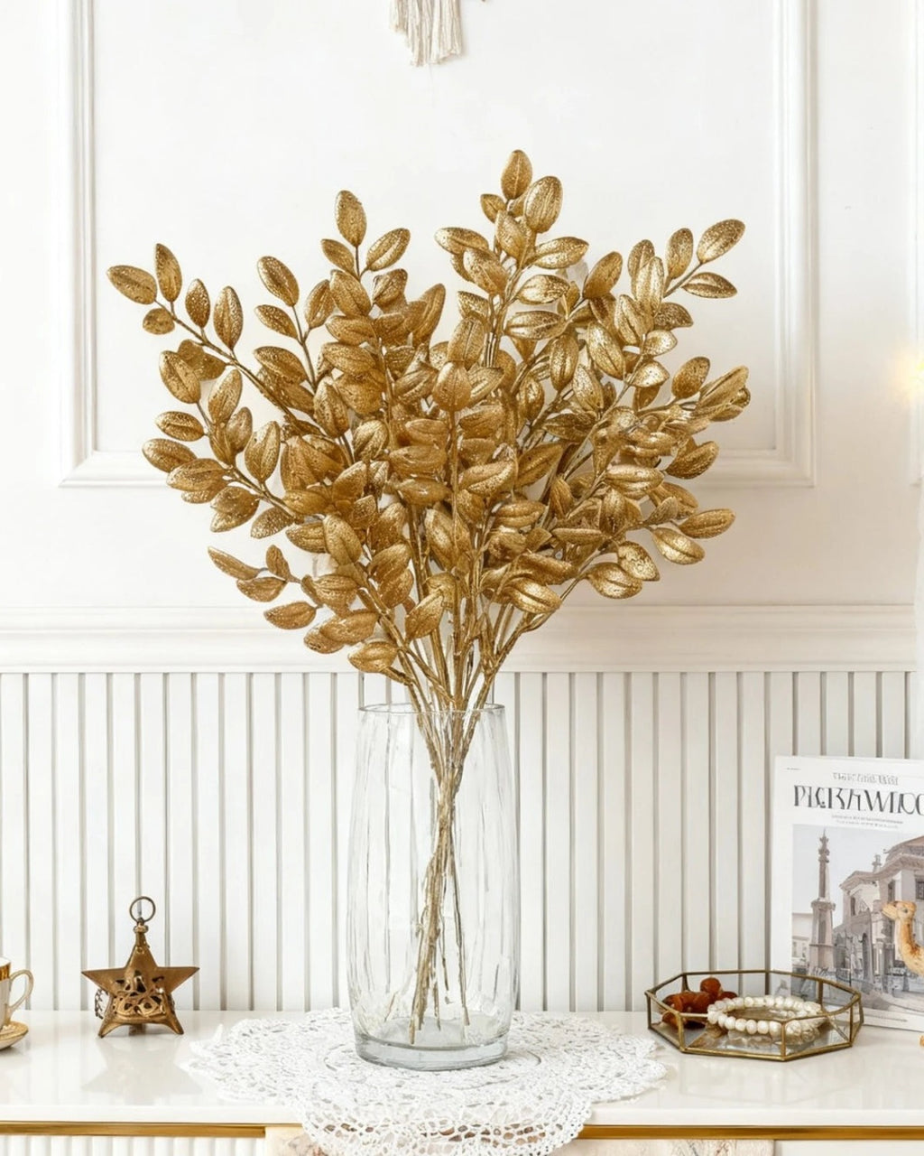 Artificial gold leaf stems in a tall glass vase against a panelled wall; no vase is included