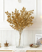 Artificial gold leaf stems in a tall glass vase against a panelled wall; no vase is included