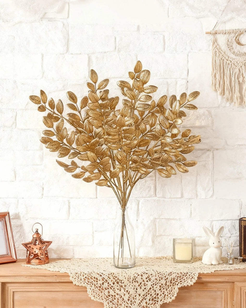Artificial gold leaf stems in a vase against a white brick wall; no vase is included