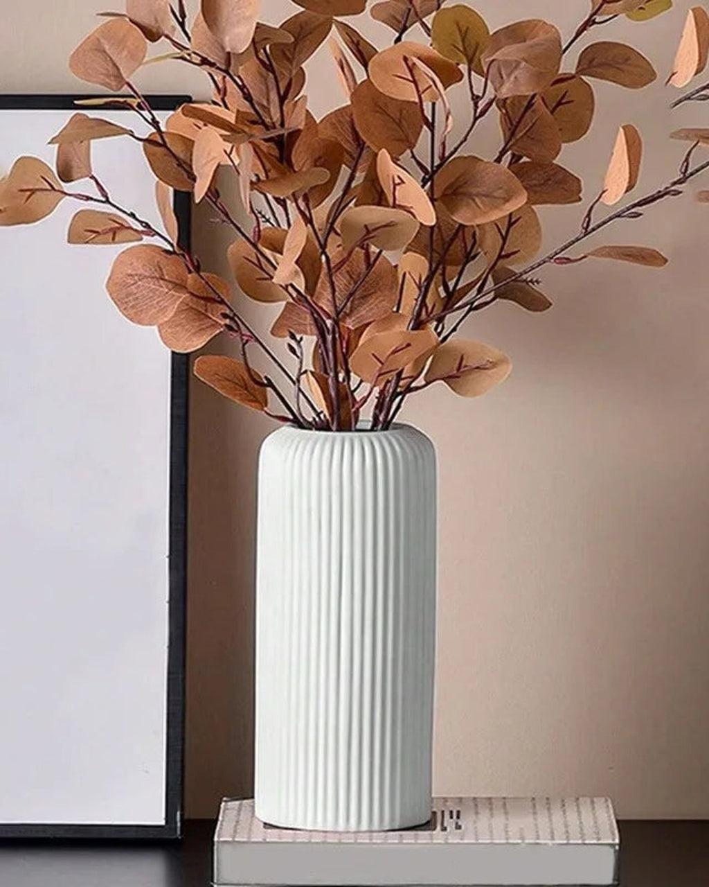 A white striped cylinder vase holding eucalyptus beside a framed print; stems not included
