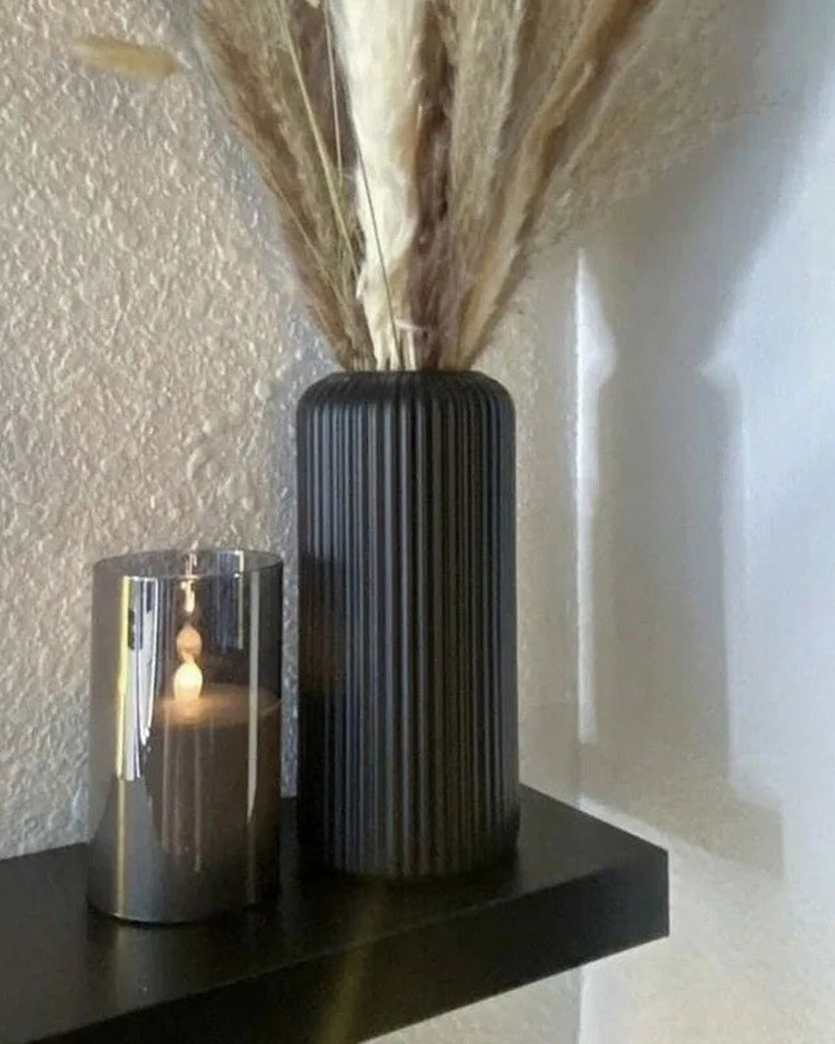 A black striped cylinder vase holding pampas on a shelf beside a candle; stems and candle not included