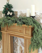 An artificial cedar garland draped over a wooden fireplace mantel; small tree and candles not included
