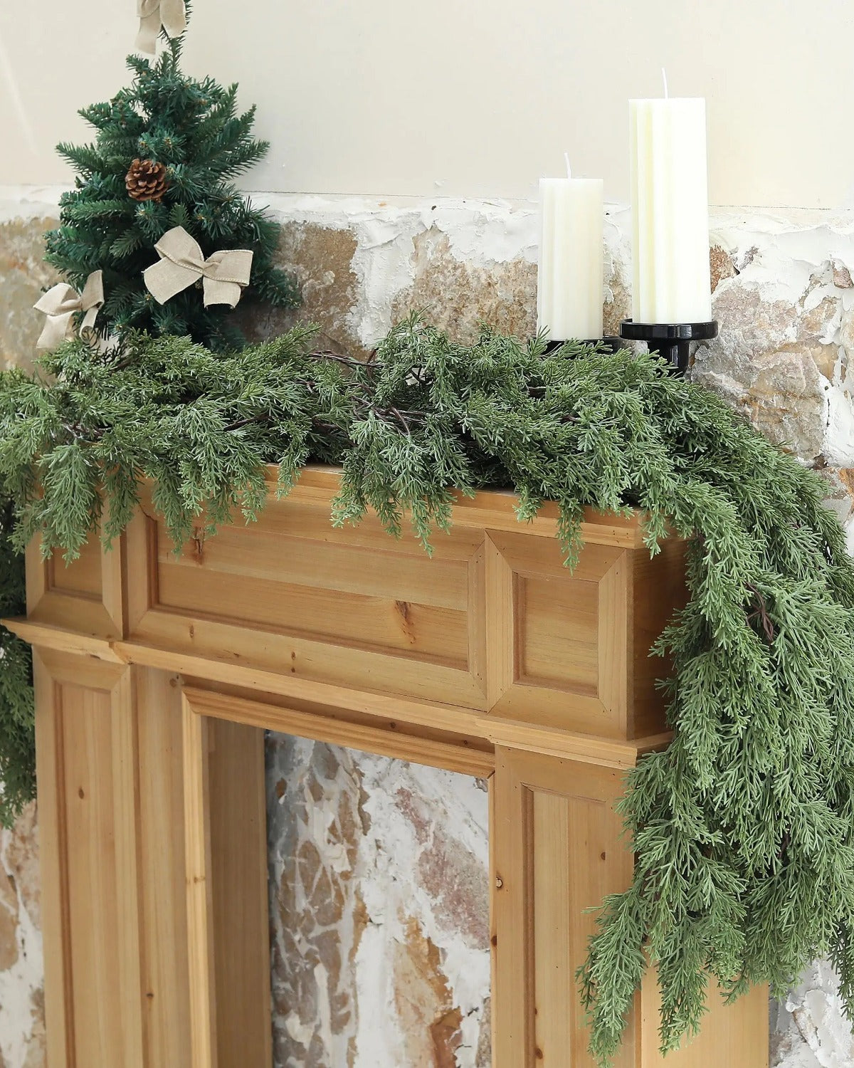 An artificial cedar garland draped over a wooden fireplace mantel; small tree and candles not included