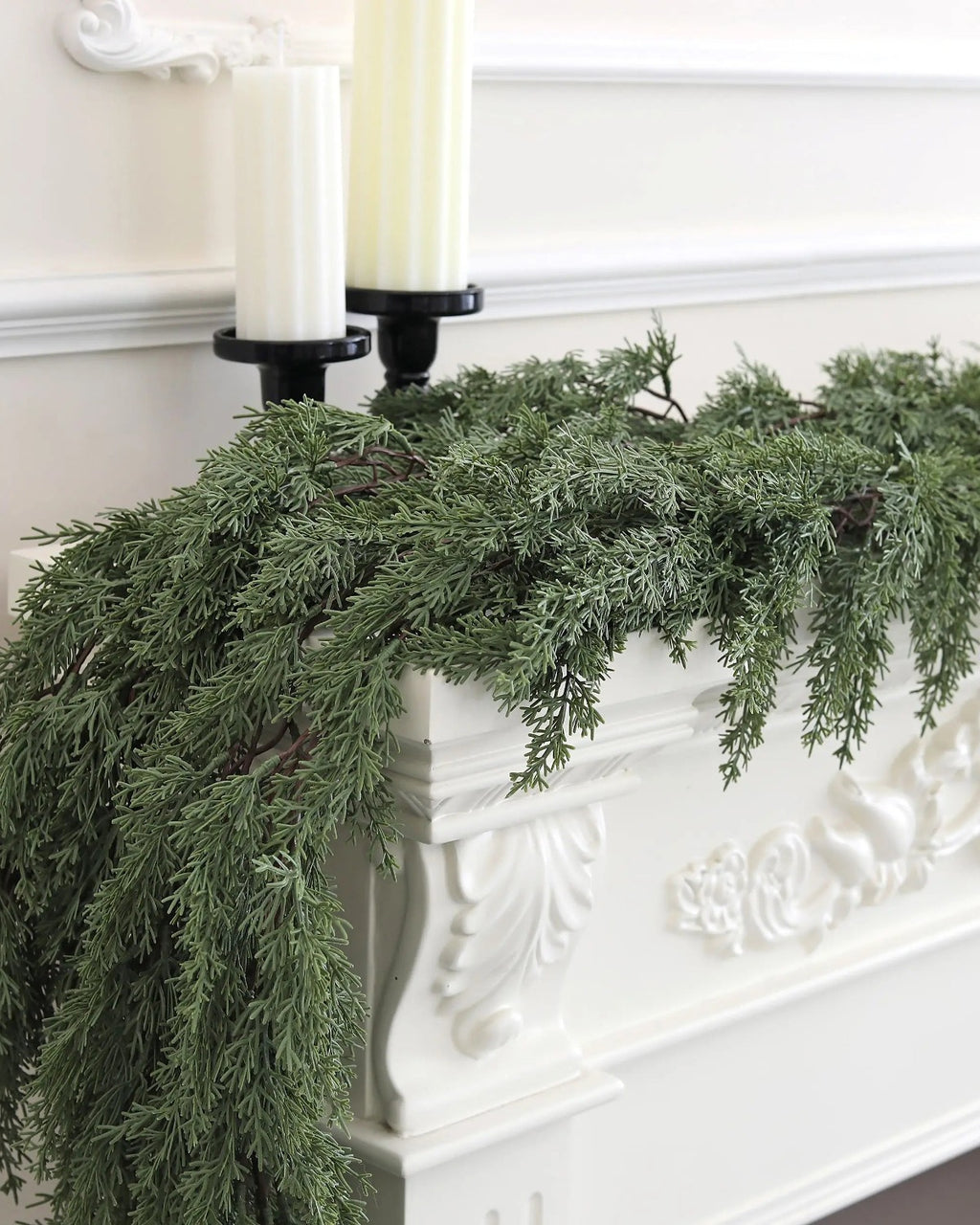 An artificial cedar garland trailing over a white carved mantel under two candles