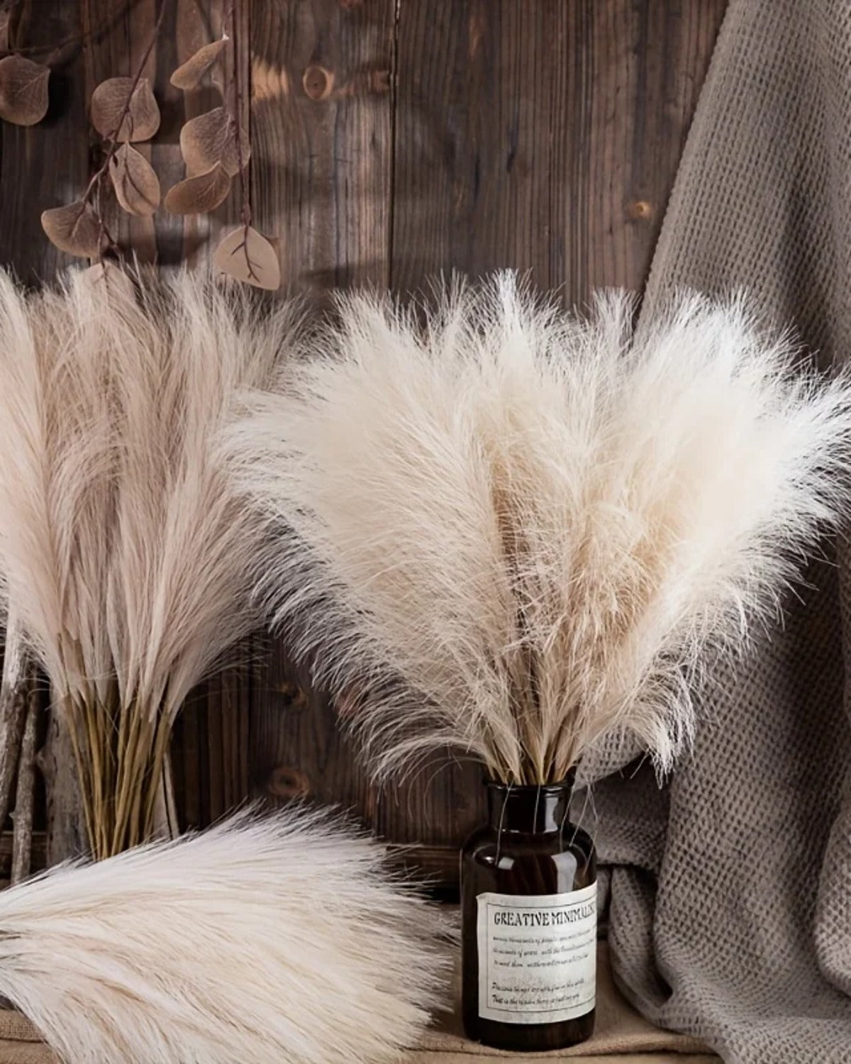 Artificial pampas stems in a dark glass bottle against a wooden wall, with more stems laid beside it