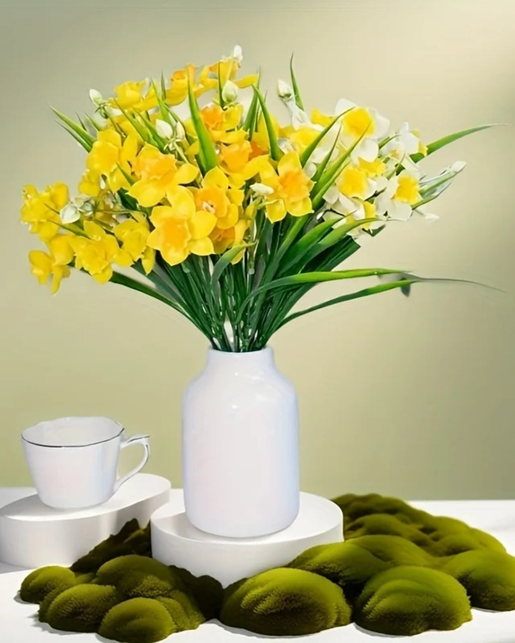 A bunch of daffodils in a white vase beside a teacup, with moss laid around the base