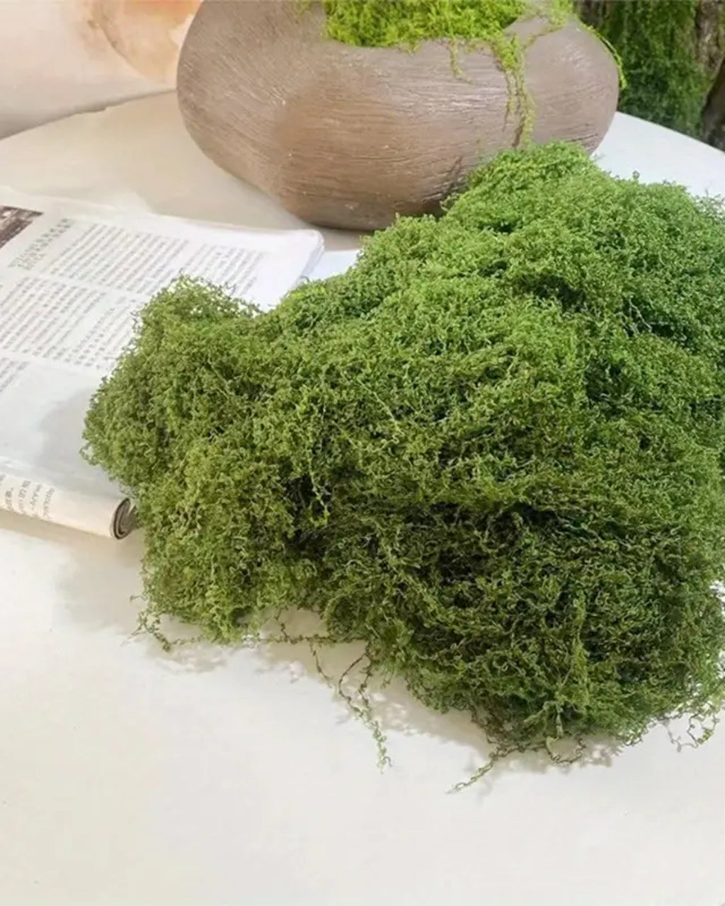 A piece of the moss mat on a table beside an open book and a wooden vessel