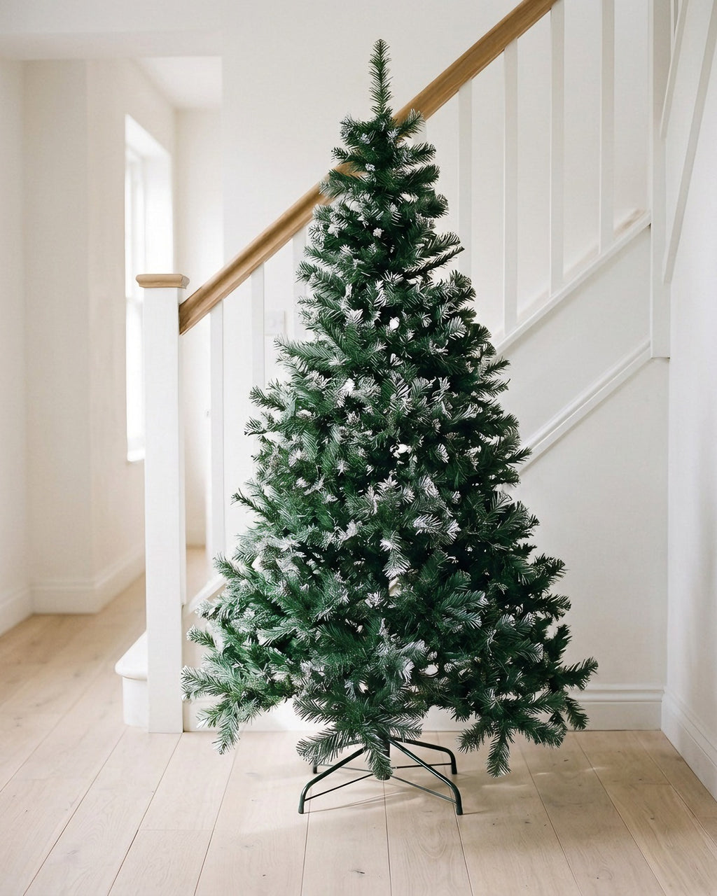 The green with frosted tips tree, undecorated, on its dark folding stand at the foot of a staircase