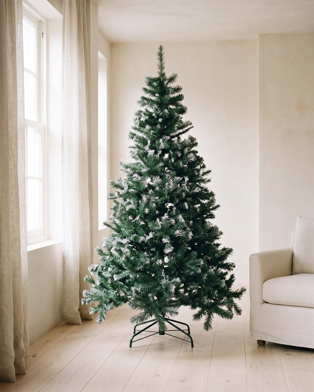 The green with frosted tips tree, undecorated, in a living room corner beside a window and an armchair