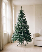 The green with frosted tips tree, undecorated, in a living room corner beside a window and an armchair