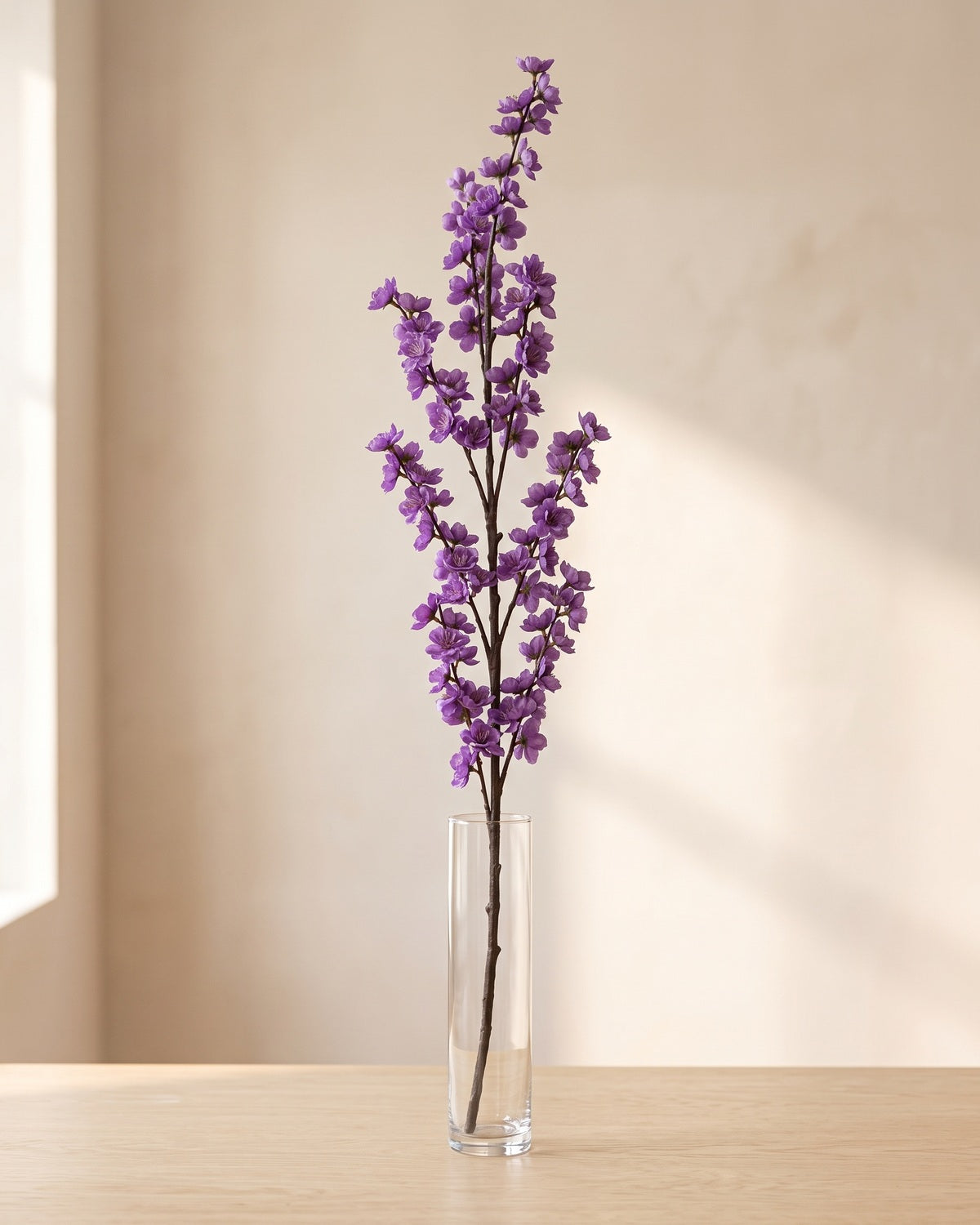One artificial peach blossom branch in a plain glass vase against a plaster wall