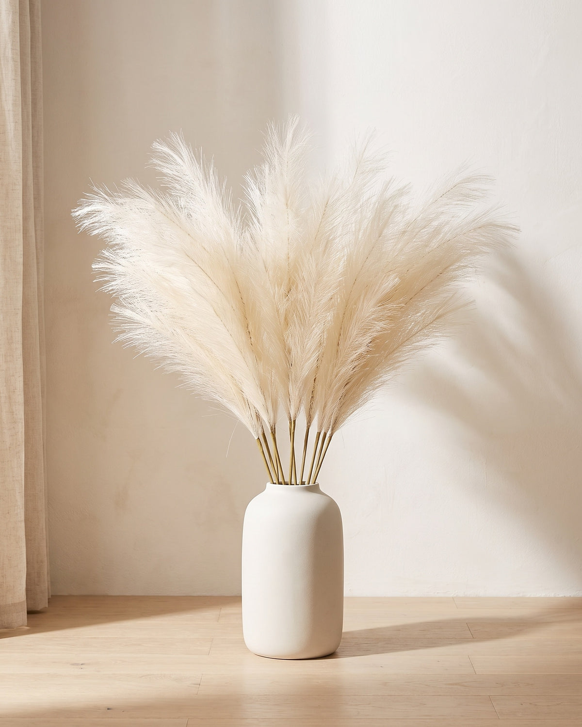 Ten artificial pampas grass stems in a plain off-white floor vase against a warm plaster wall