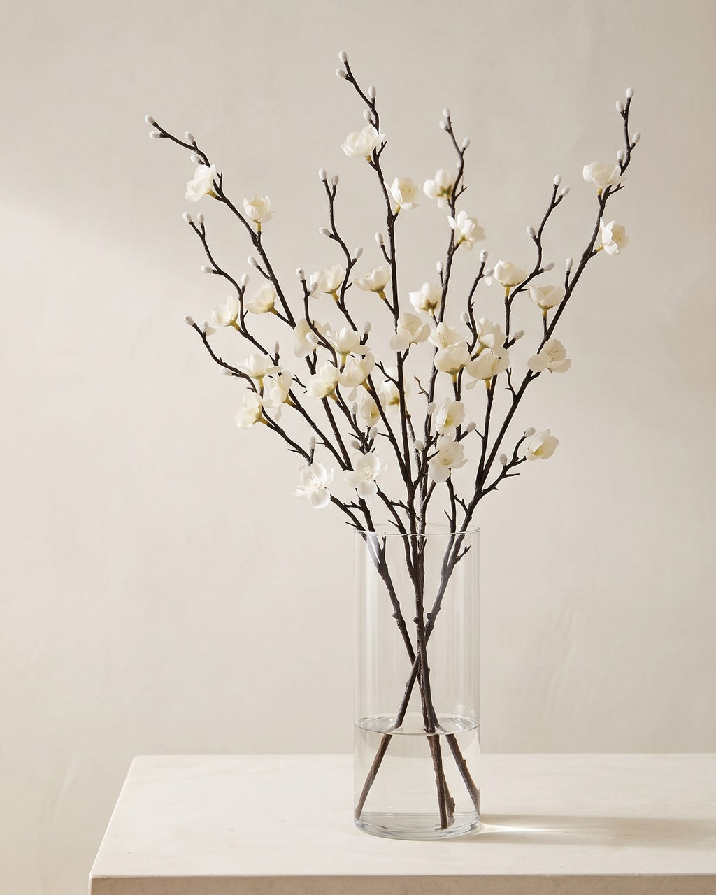 Five artificial white plum blossom stems in a plain clear glass vase on a pale surface. Vase not included.
