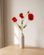 Three artificial red poppy stems in a slim off-white bud vase on a pale oak tabletop