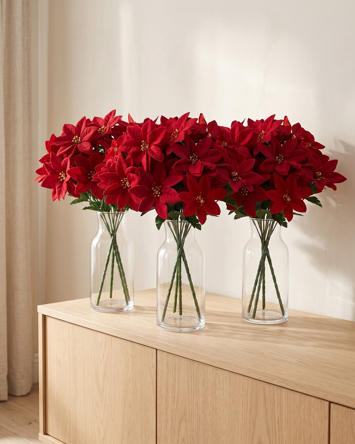 Red artificial poinsettia bouquets in three clear glass vases on a sideboard; vases not included