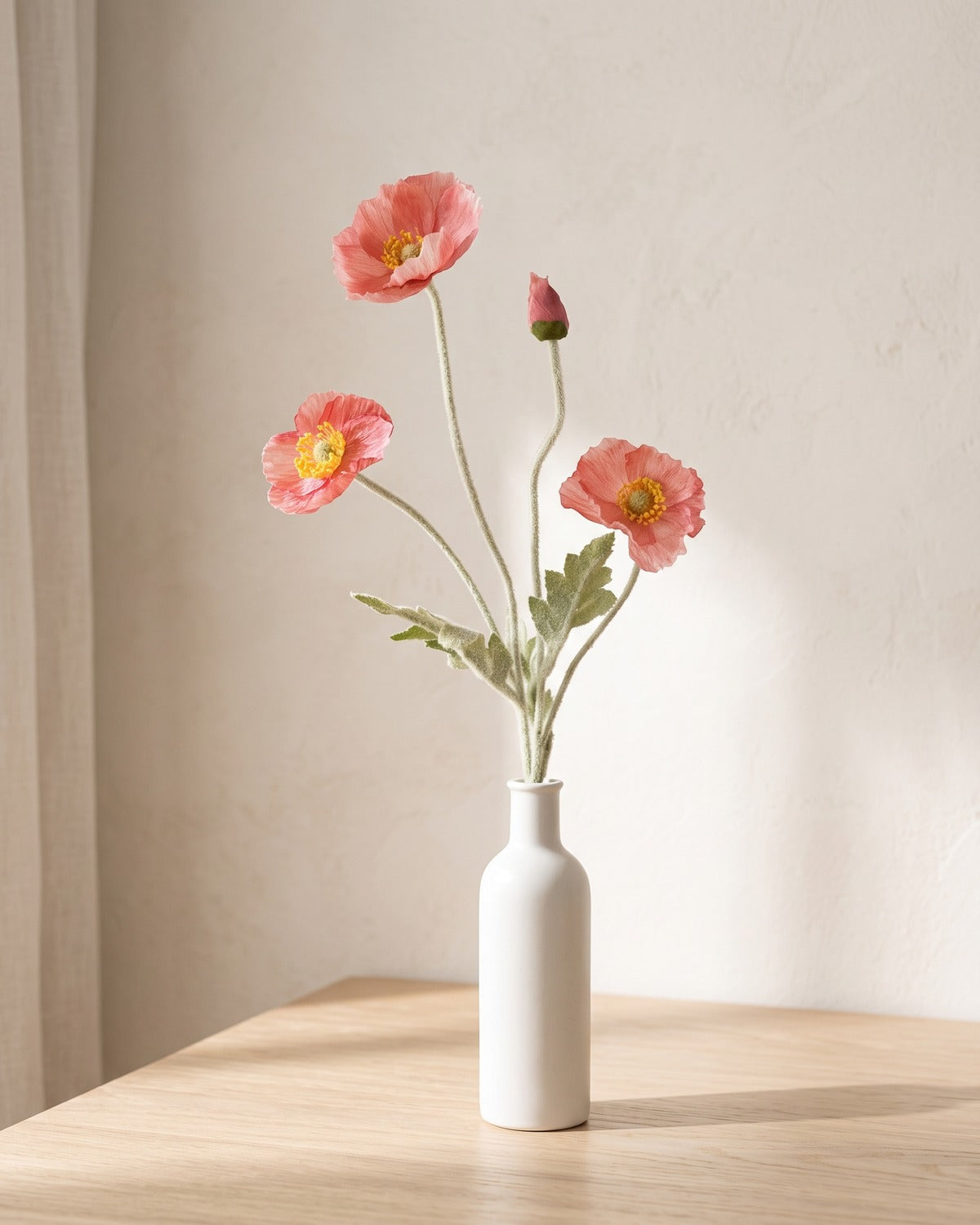 A dark pink artificial poppy stem in a plain white bottle vase on a pale oak table; vase not included