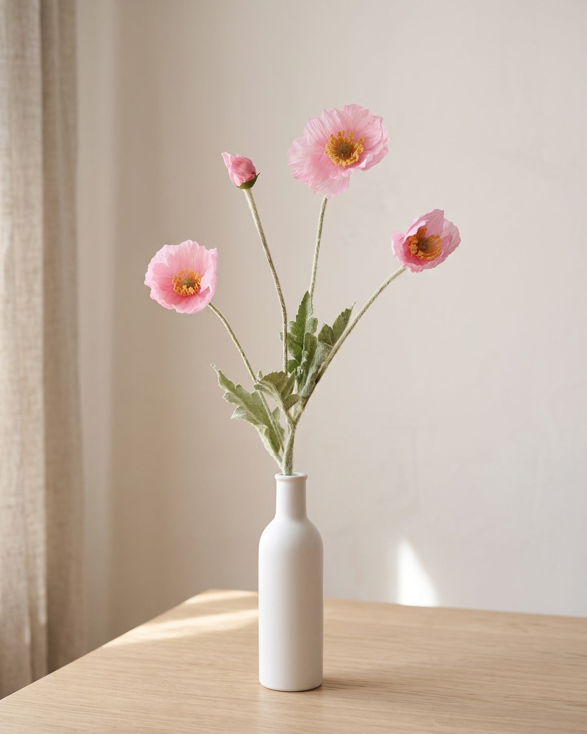 A fuchsia artificial poppy stem in a plain white bottle vase on a pale oak table; vase not included