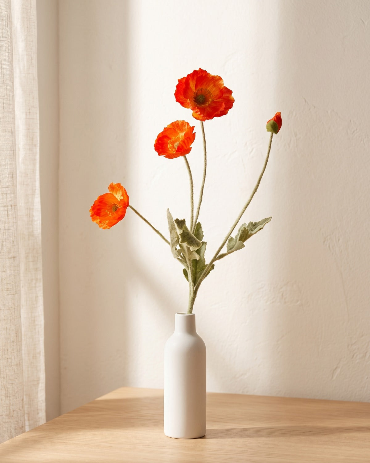 A red orange artificial poppy stem in a plain white bottle vase on a pale oak table; vase not included