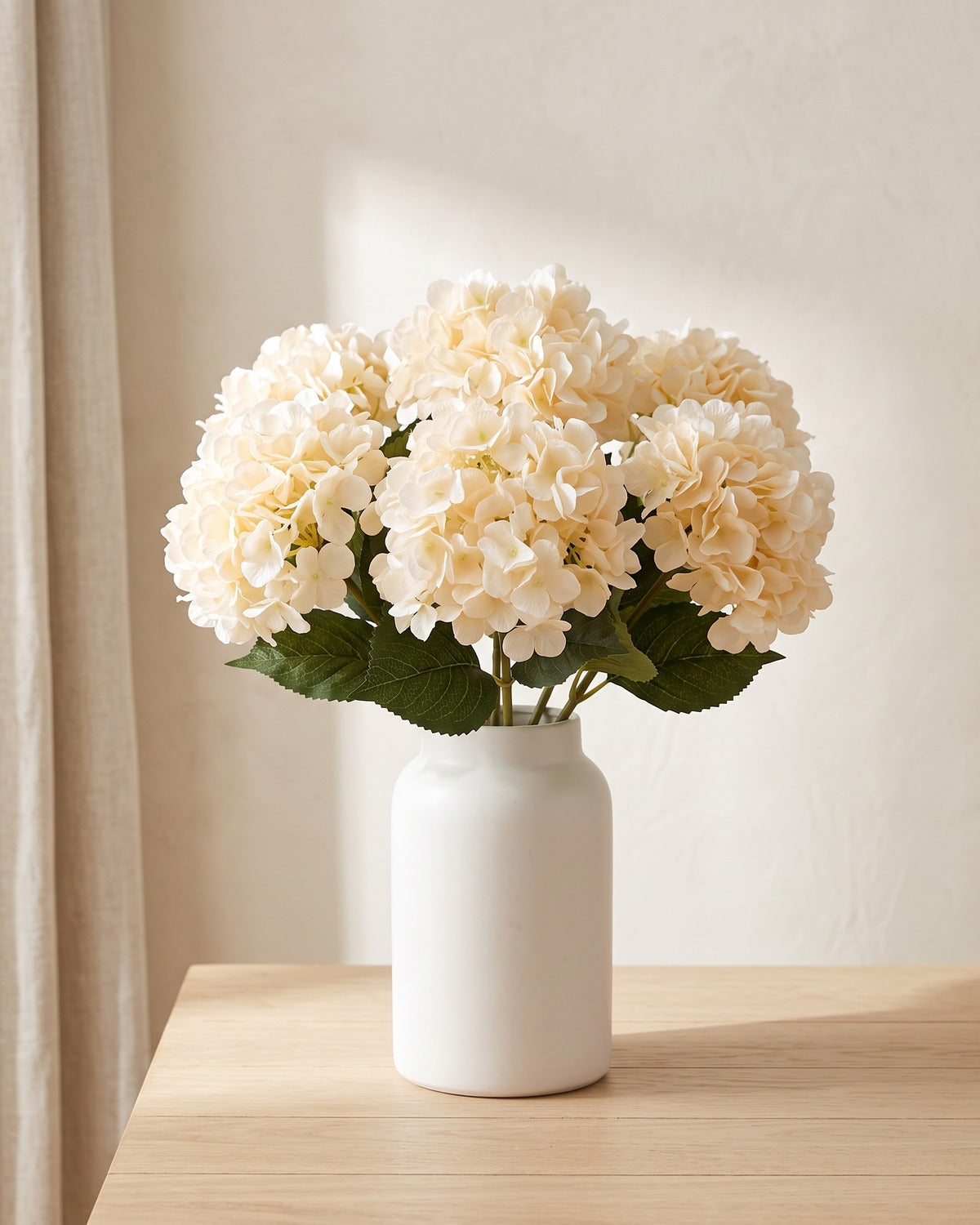 Champagne artificial hydrangea flowers standing in a plain white vase on a pale oak table; no vase is included