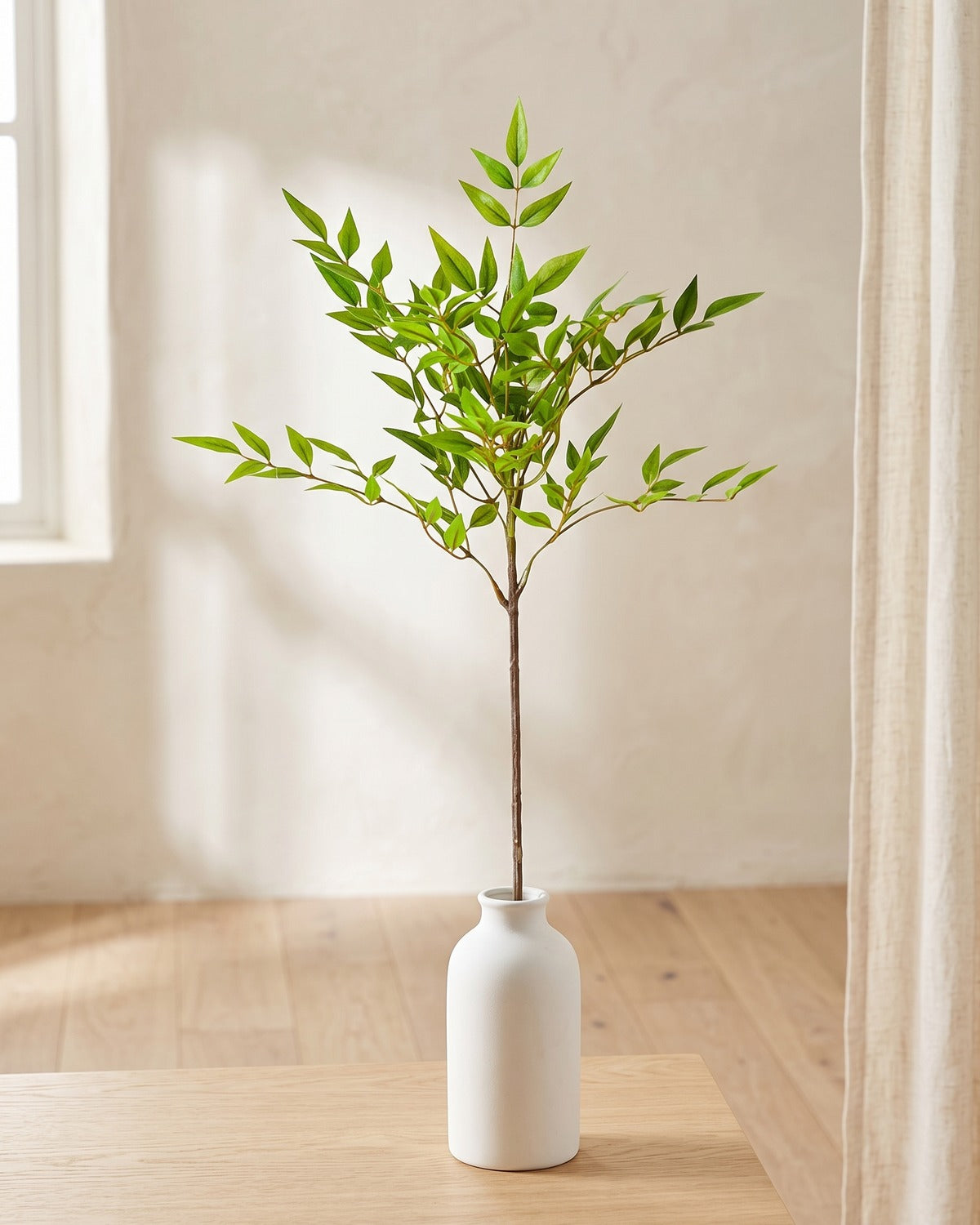 Fine leaf artificial tree branch, 1 stem, 30 in, standing in a plain white vase on a pale oak table; no vase is included