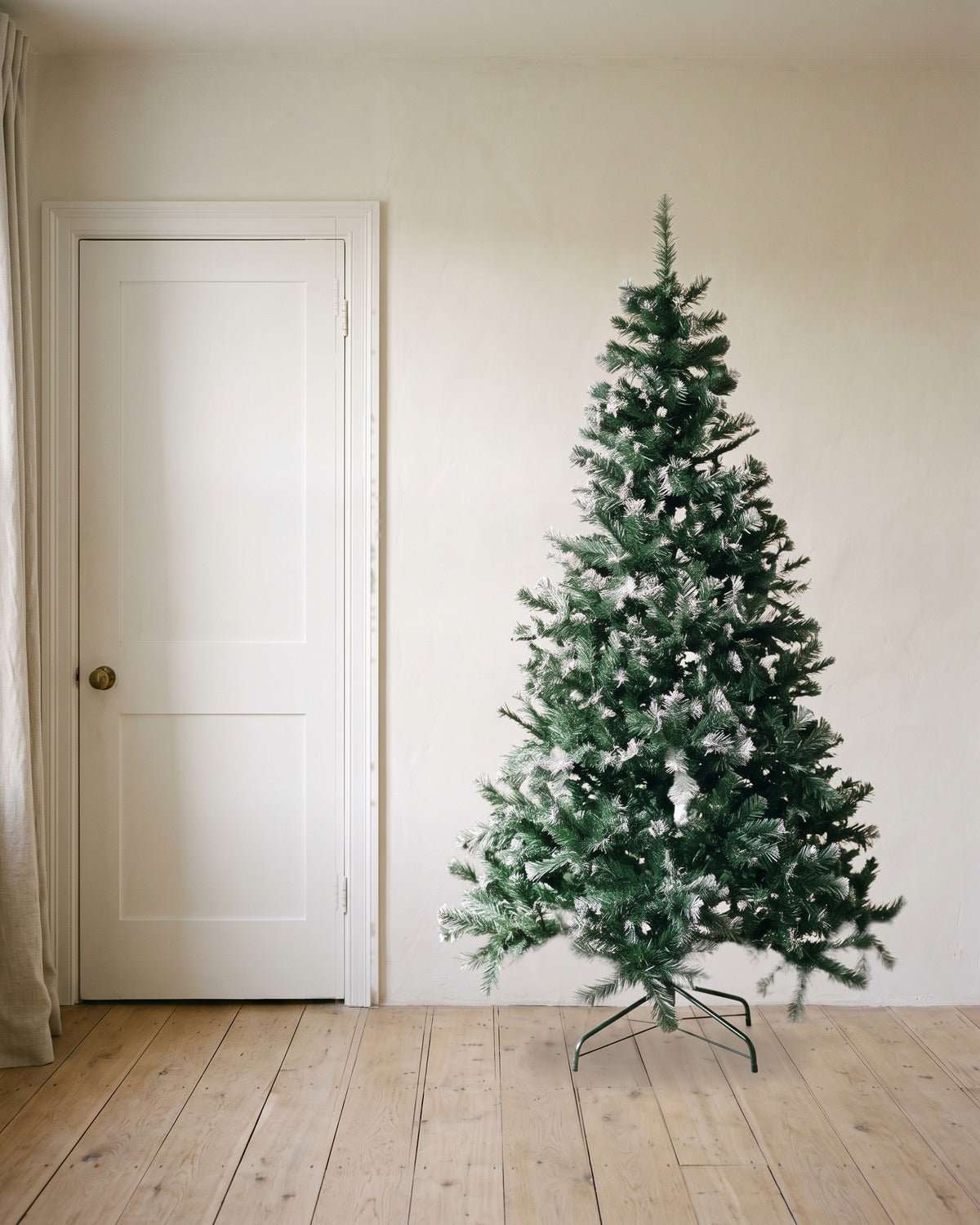 A green artificial Christmas tree with frosted tips at 7 ft on its folding stand, beside an 80 in door to show its height
