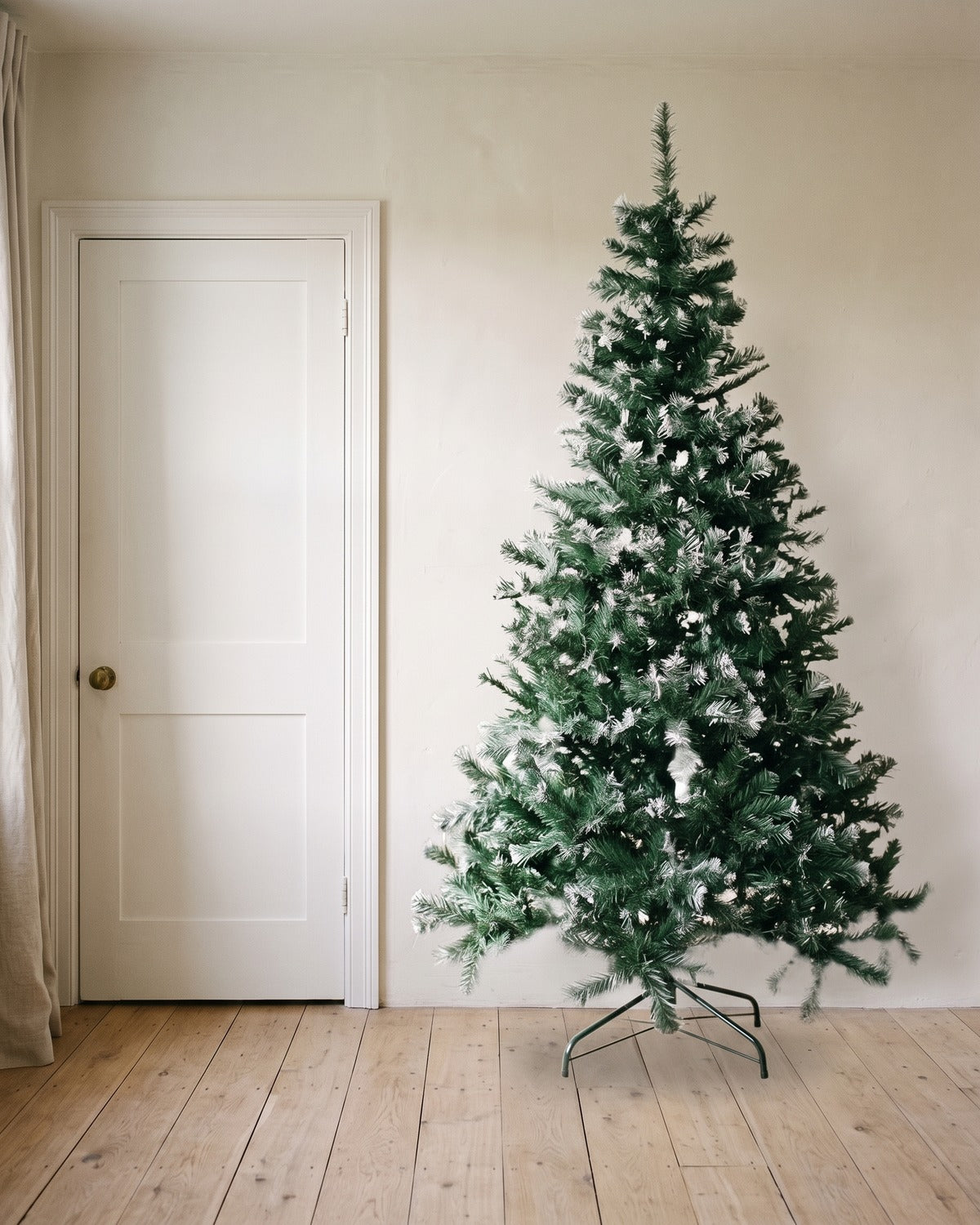 A green artificial Christmas tree with frosted tips at 8 ft on its folding stand, beside an 80 in door to show its height