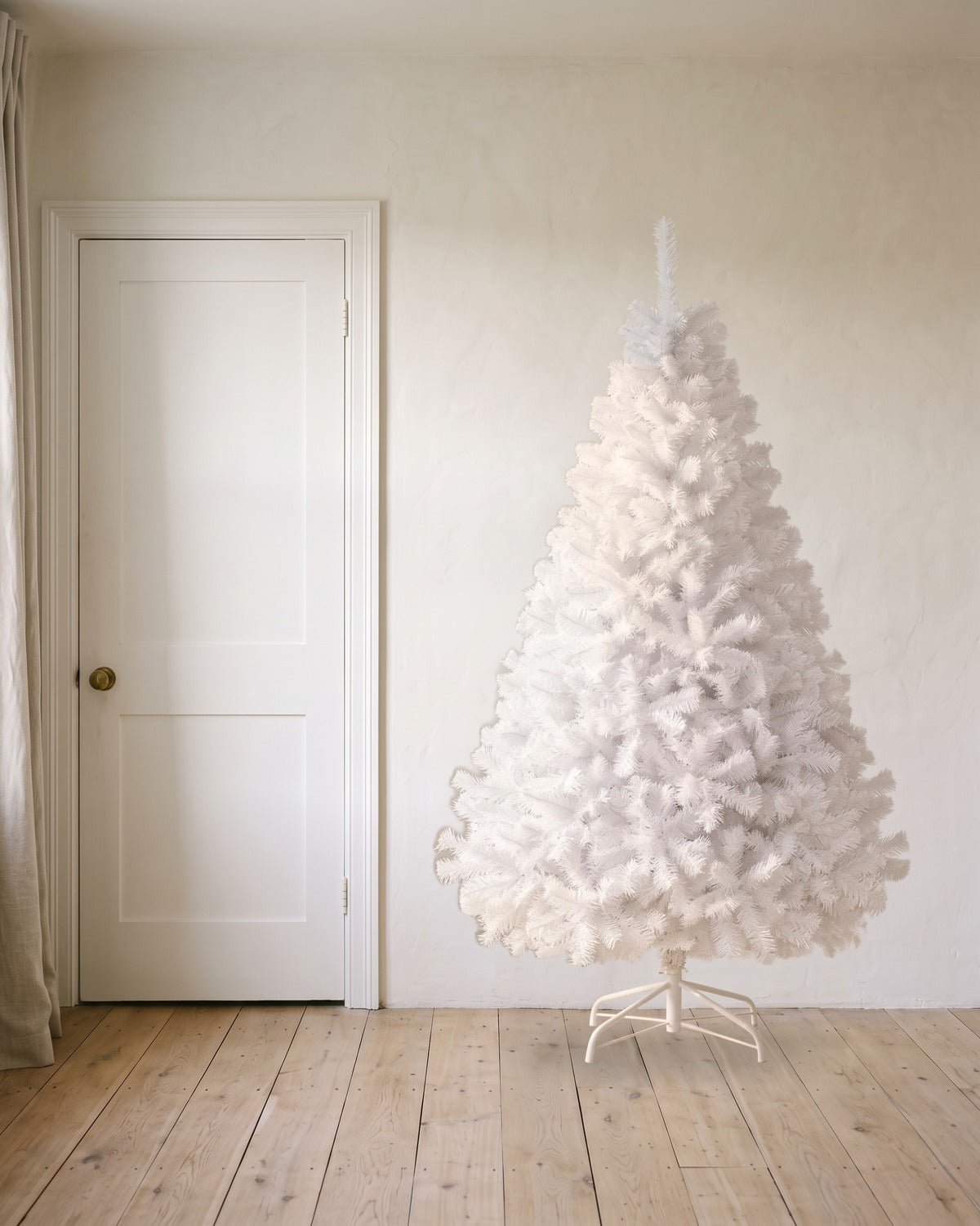 A white artificial Christmas tree at 7 ft on its folding stand, beside an 80 in door to show its height