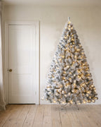 A pre-lit snow flocked Christmas tree at 7.5 ft, its lights on, on its stand beside an 80 in door to show its height
