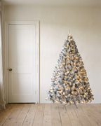 A pre-lit snow flocked Christmas tree at 6 ft, its lights on, on its stand beside an 80 in door to show its height