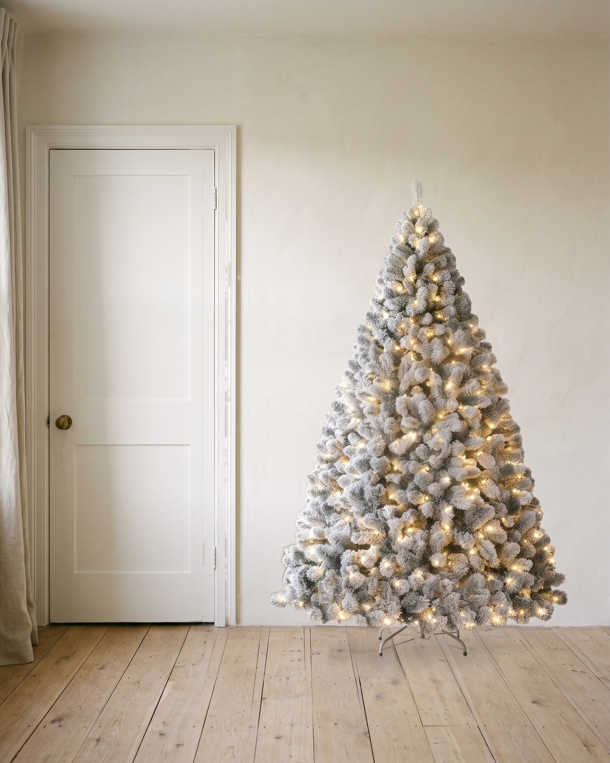 A pre-lit snow flocked Christmas tree at 6 ft, its lights on, on its stand beside an 80 in door to show its height